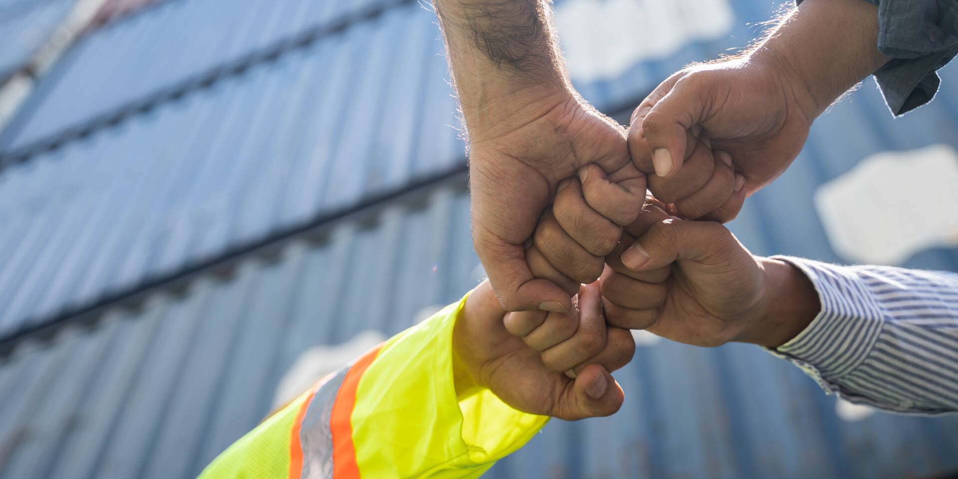Two construction workers in safety vests and striped shirt perform a fist bump, symbolizing teamwork and partnership.