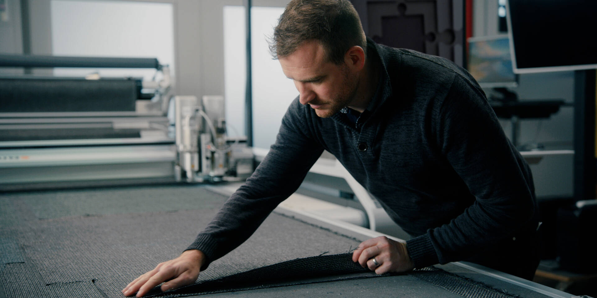 A man inspects a roll of dark fabric on a cutting table next to a Zünd cutting system.