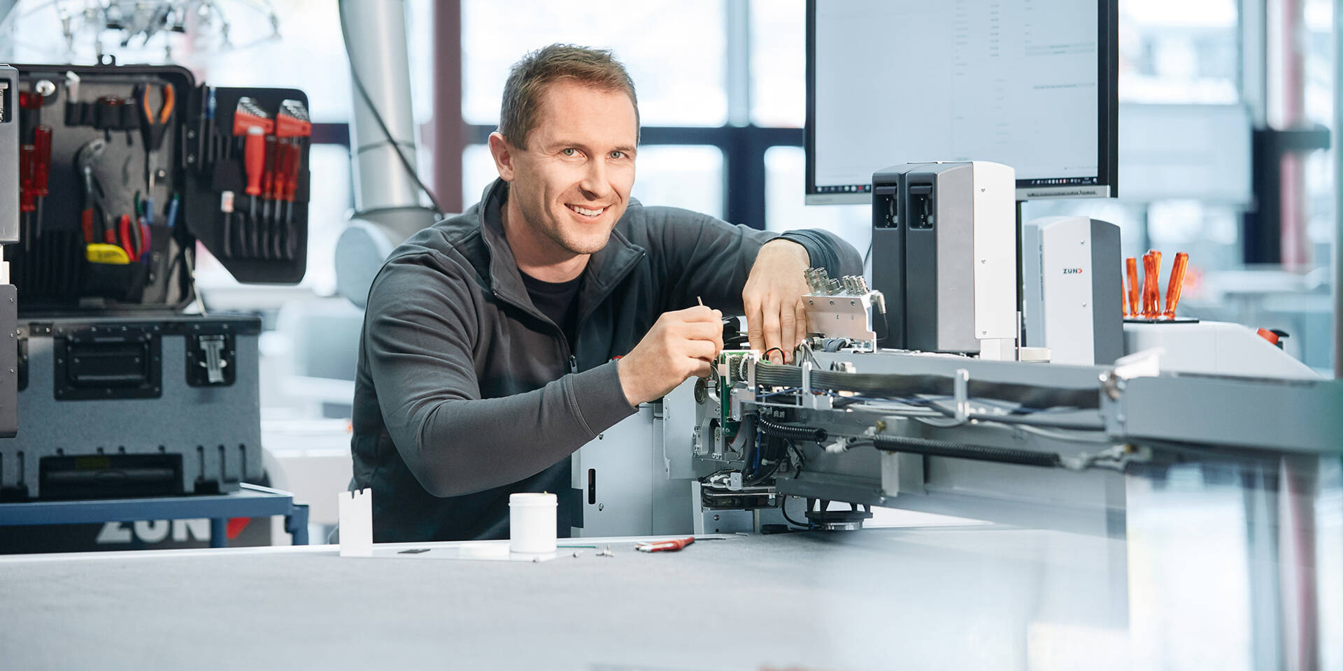 A smiling technician works on a sophisticated Zünd cutting machine with tools and equipment visible in the background.