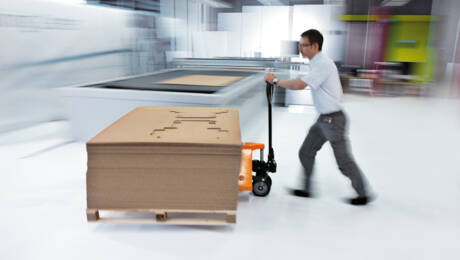 A man pushes a pallet jack loaded with a stack of die-cut cardboard sheets in a modern production facility. A man pushes a pallet jack loaded with a stack of die-cut cardboard sheets in a modern production facility.