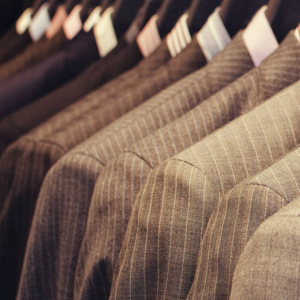Close-up of a row of men's suits and jackets with subtle pinstripe and textured patterns, hanging on display. Close-up of a row of men's suits and jackets with subtle pinstripe and textured patterns, hanging on display.