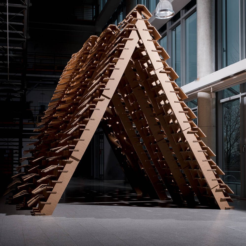 Struttura architettonica a forma di tenda, realizzata con pannelli di cartone tagliati con precisione, in un ambiente interno. Struttura architettonica a forma di tenda, realizzata con pannelli di cartone tagliati con precisione, in un ambiente interno.