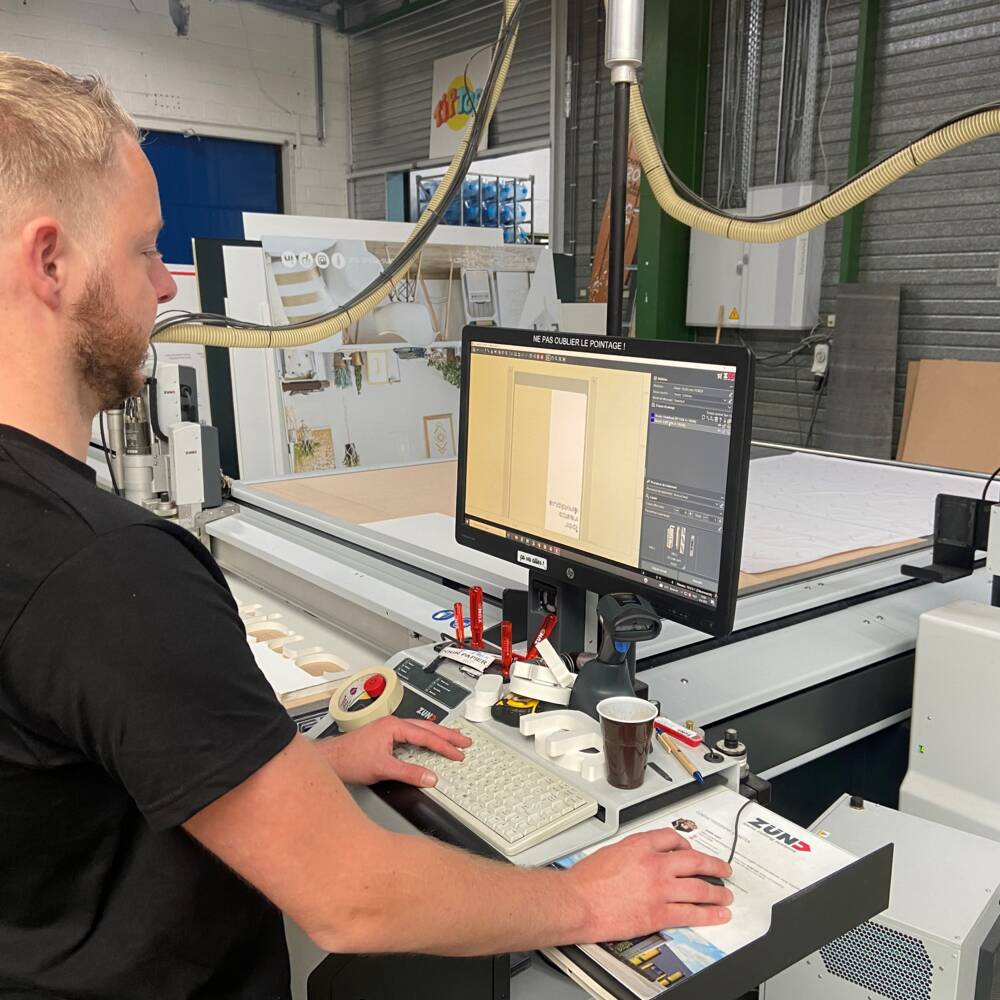 Man in zwart shirt kijkt naar Zünd snijplotter met computer die ontwerp toont, in een werkplaatsomgeving. Man in zwart shirt kijkt naar Zünd snijplotter met computer die ontwerp toont, in een werkplaatsomgeving.