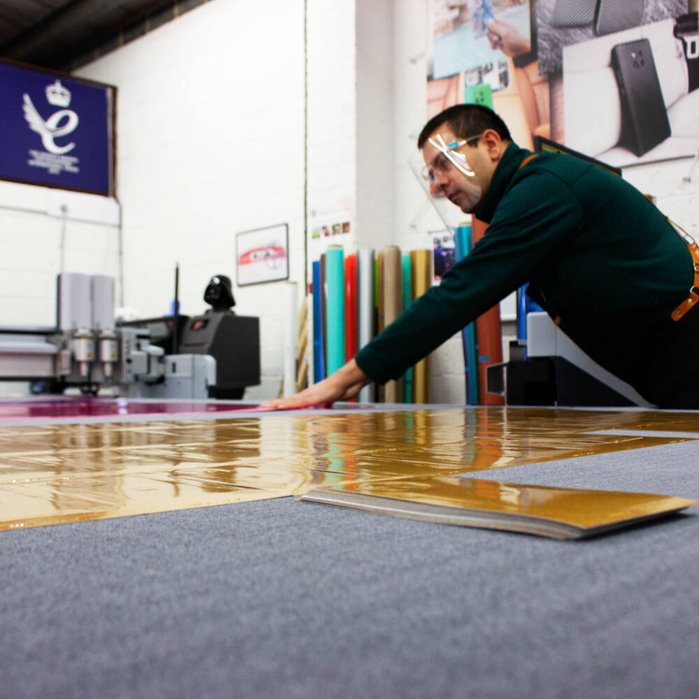 A man wearing a face shield works with large sheets of gold vinyl next to a Zünd cutting machine in a workshop. A man wearing a face shield works with large sheets of gold vinyl next to a Zünd cutting machine in a workshop.