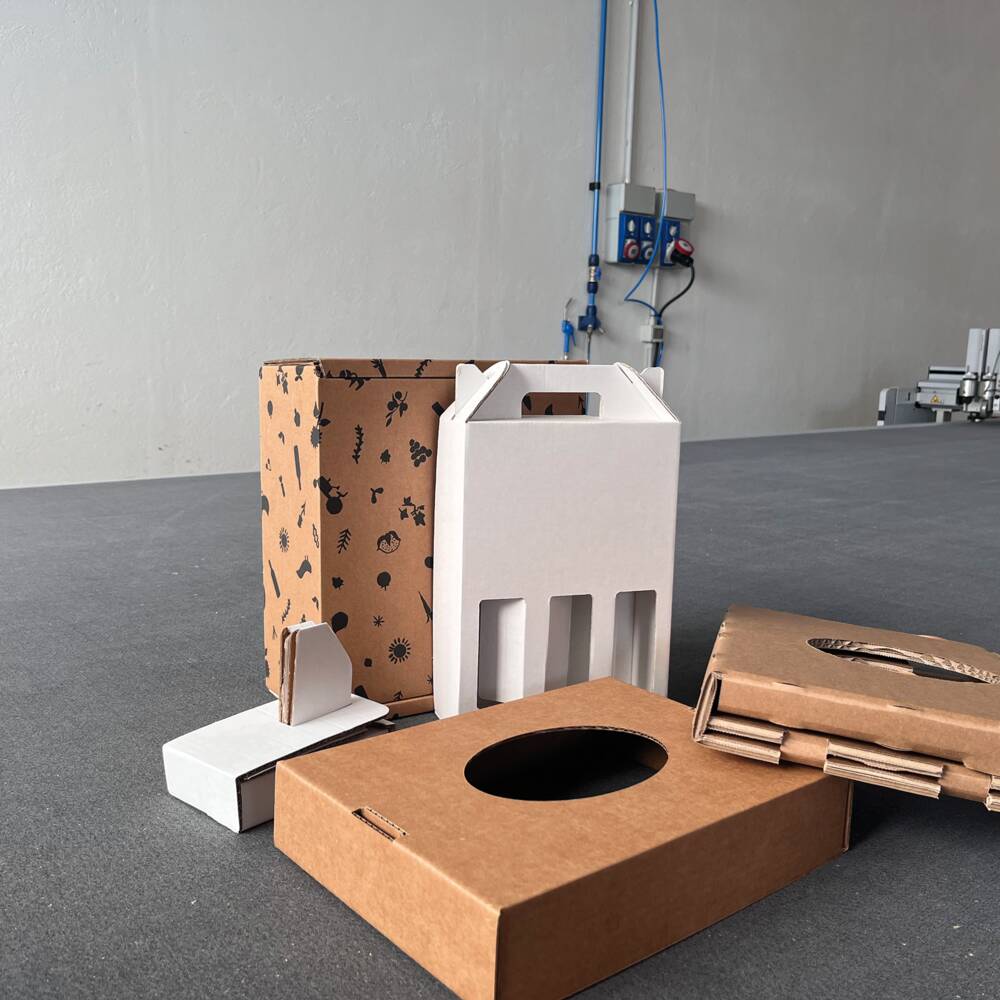 Scatole di cartone fustellate, una bianca con manico e una marrone con motivi neri, su un tavolo da taglio. Scatole di cartone fustellate, una bianca con manico e una marrone con motivi neri, su un tavolo da taglio.