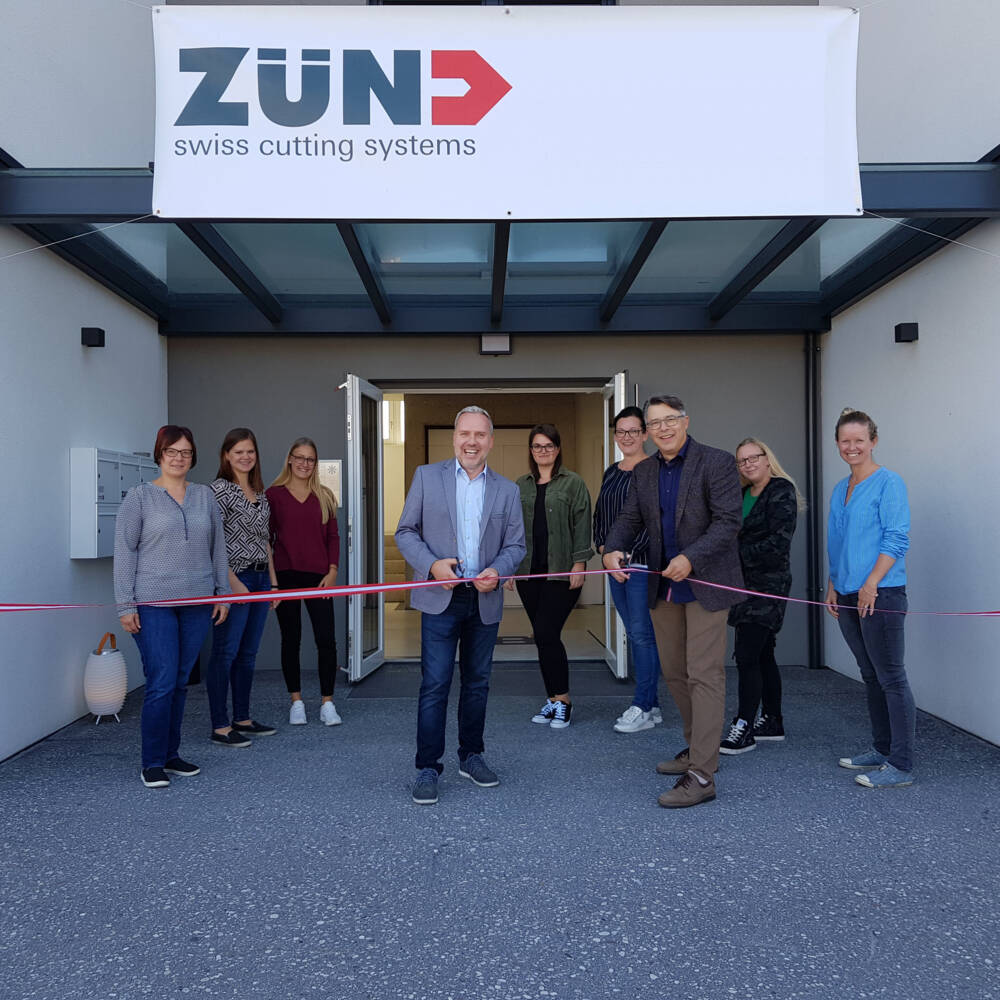 Une équipe souriante de Zünd Systemtechnik coupe le ruban lors d'une cérémonie d'ouverture, devant un bâtiment moderne. Une équipe souriante de Zünd Systemtechnik coupe le ruban lors d'une cérémonie d'ouverture, devant un bâtiment moderne.
