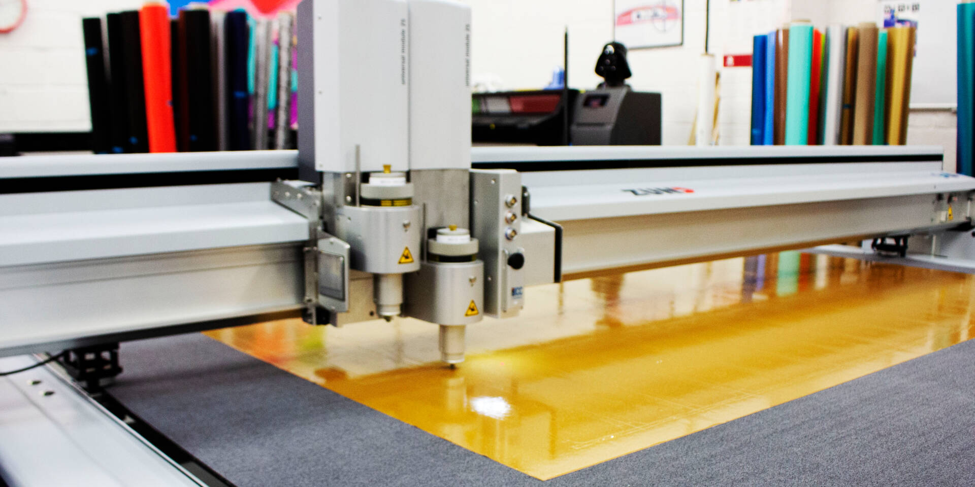 Zünd G3 L-2500 digital cutter precisely scores a large sheet of reflective orange material on a work surface.