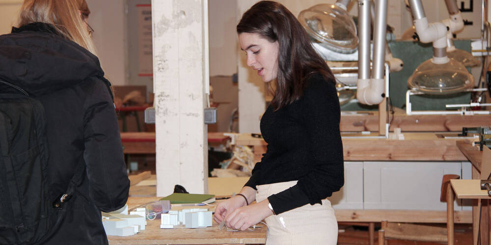 Two people examine architectural models on a wooden table in a workshop setting, with Zünd cutting equipment visible in the background.