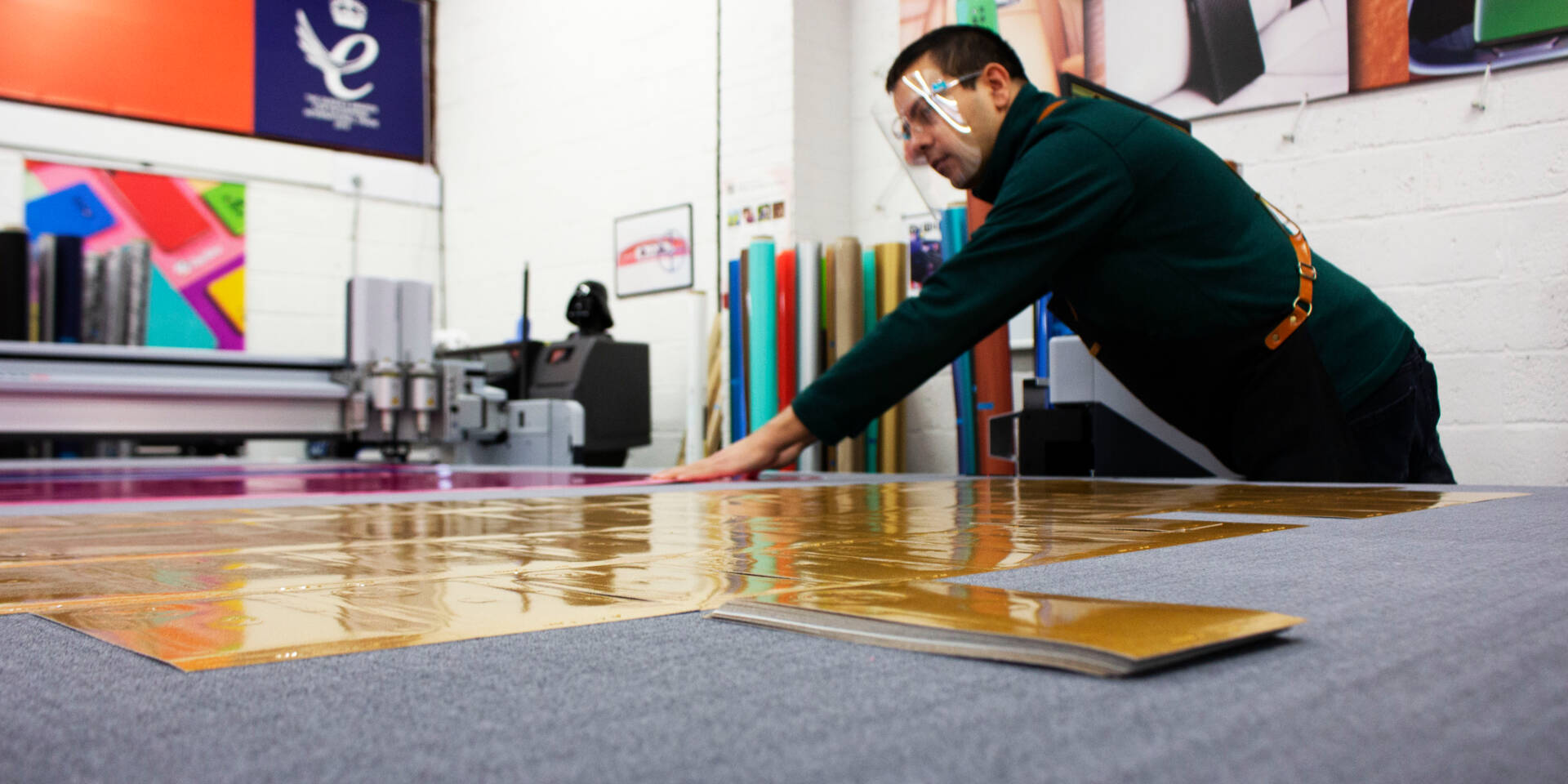 A man wearing a face shield works with large sheets of gold vinyl next to a Zünd cutting machine in a workshop.