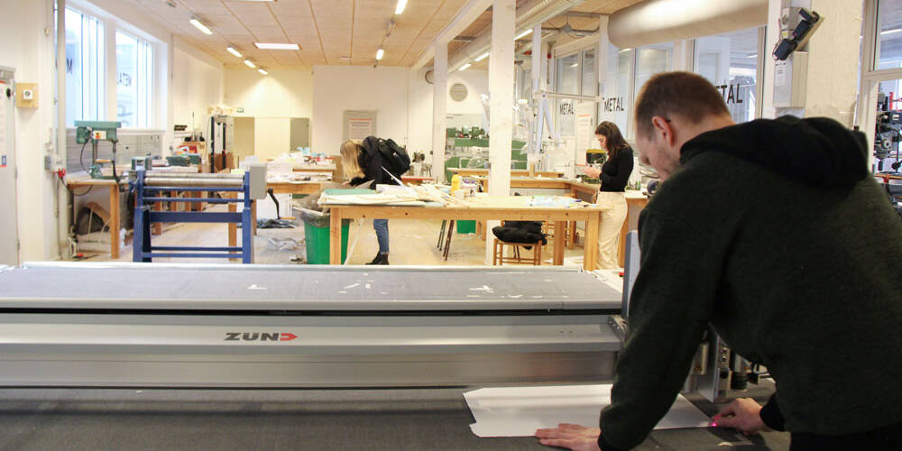 A person operates a Zünd cutting machine in a workshop with other people working at tables in the background.