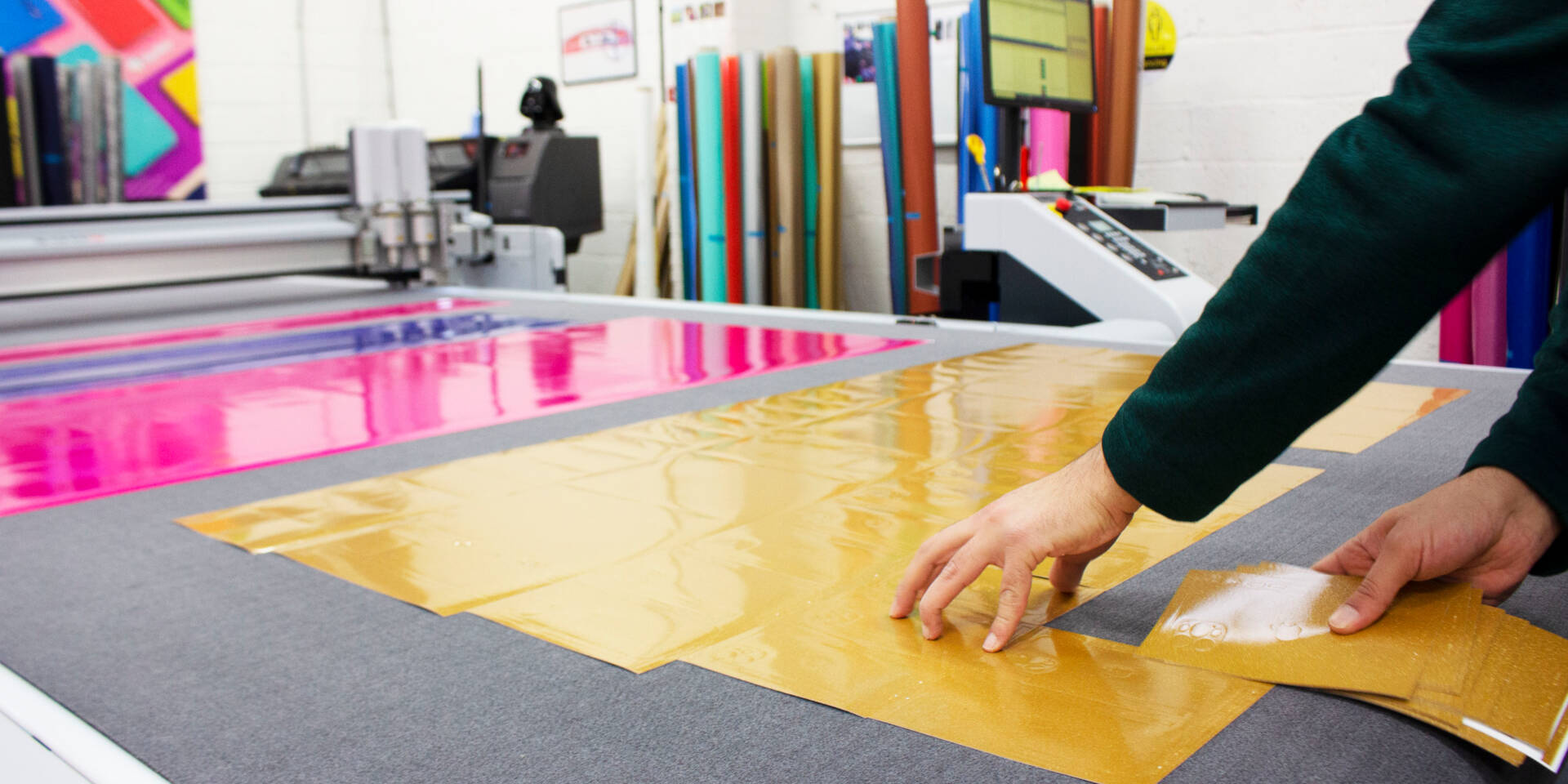A person's hands place gold material onto a cutting mat next to pink material, with a Zünd cutter in the background.