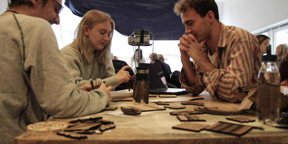 Students working on woodcraft projects at a table, with a laptop and projector in the background.