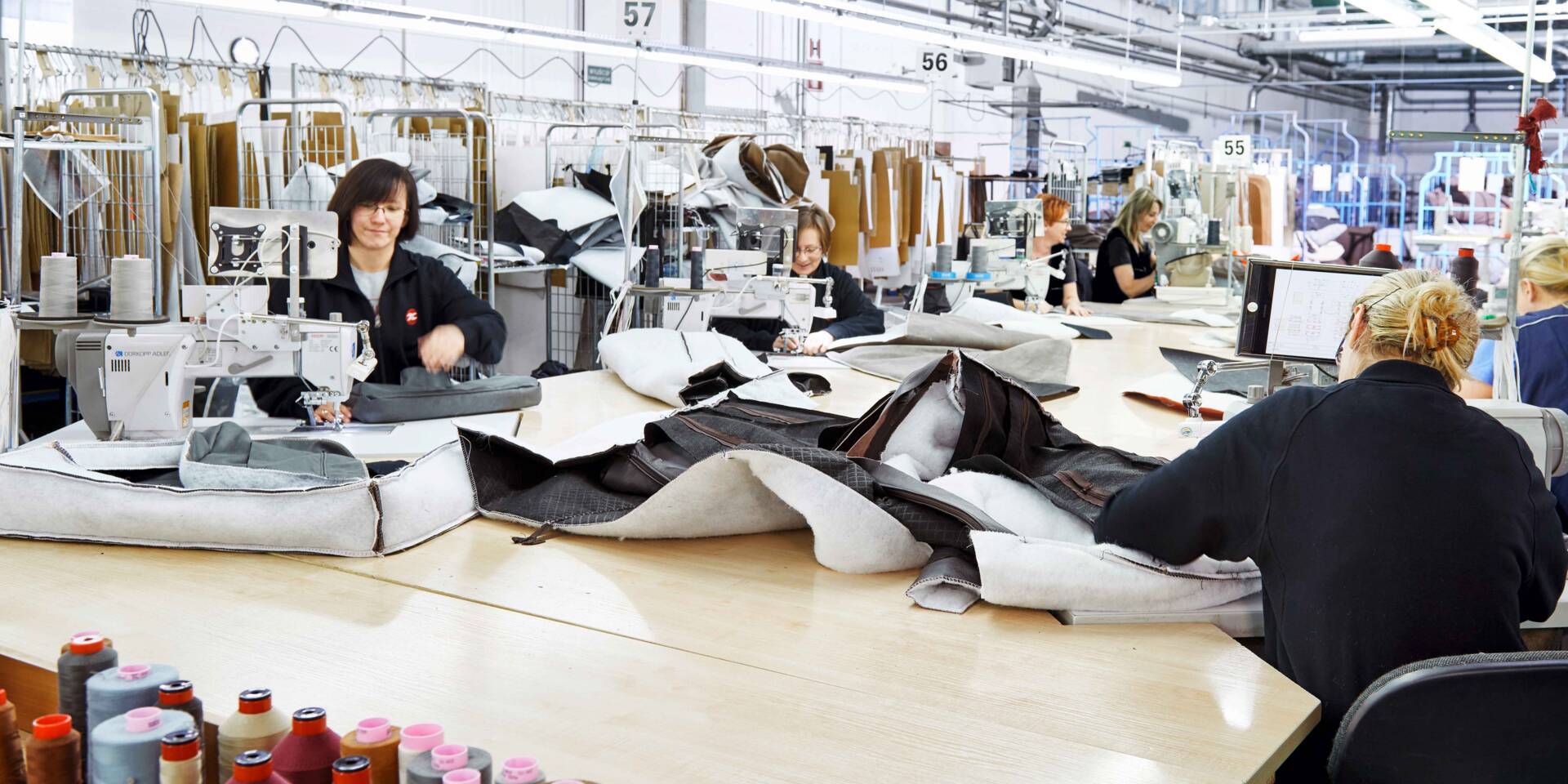 Operai in una fabbrica di abbigliamento lavorano a macchine da cucire industriali su un tavolo di legno, con materiali e fili sparsi.