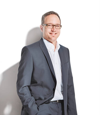 Ritratto di un uomo sorridente con un abito grigio scuro e una camicia bianca, su sfondo bianco con ombra.