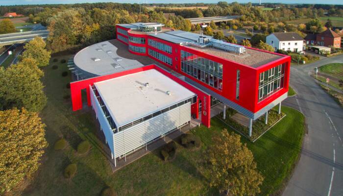 Edificio aziendale moderno con facciate rosse e bianche, circondato da verde e alberi in autunno. Si vede un cartello 'GRUPPE'. Edificio aziendale moderno con facciate rosse e bianche, circondato da verde e alberi in autunno. Si vede un cartello 'GRUPPE'.