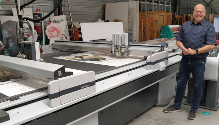 Man staat naast een Zünd D3 XL-3200 snijplotter in een werkplaats met diverse prints. Man staat naast een Zünd D3 XL-3200 snijplotter in een werkplaats met diverse prints.