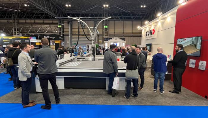 People gather around a large Zünd digital cutting system at an exhibition booth, showcasing its capabilities with various materials. People gather around a large Zünd digital cutting system at an exhibition booth, showcasing its capabilities with various materials.