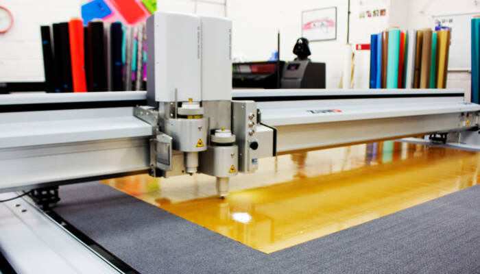 Zünd G3 L-2500 digital cutter precisely scores a large sheet of reflective orange material on a work surface. Zünd G3 L-2500 digital cutter precisely scores a large sheet of reflective orange material on a work surface.