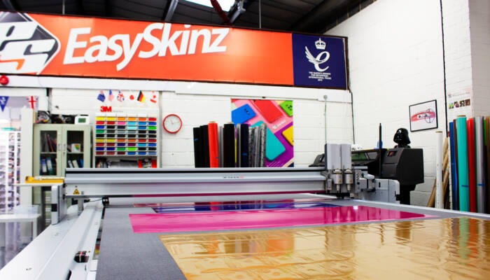 A Zünd G3 L-3200 digital cutting machine precisely cuts colorful vinyl sheets in a workshop environment. Features EasySkinz branding. A Zünd G3 L-3200 digital cutting machine precisely cuts colorful vinyl sheets in a workshop environment. Features EasySkinz branding.