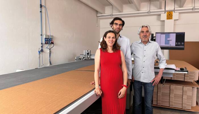 Matilde, Giulio e Danilo Tosato (da sinistra). Tre persone in piedi davanti a un banco da lavoro con materiale da imballaggio e una macchina da taglio Zünd, in un ambiente industriale.