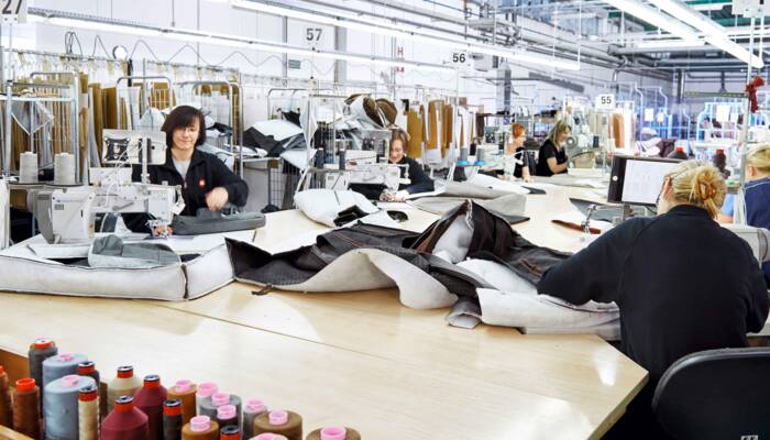 Operai in una fabbrica di abbigliamento lavorano a macchine da cucire industriali su un tavolo di legno, con materiali e fili sparsi. Operai in una fabbrica di abbigliamento lavorano a macchine da cucire industriali su un tavolo di legno, con materiali e fili sparsi.