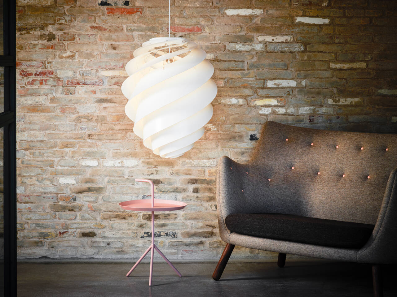 A white spiral pendant light hangs above a small pink side table next to a tufted grey armchair against a brick wall. A white spiral pendant light hangs above a small pink side table next to a tufted grey armchair against a brick wall.