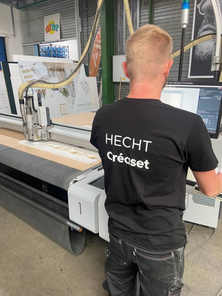Man in zwart T-shirt met 'HECHT Créaset' op de rug, bedient een Zünd snijmachine in een werkplaats.